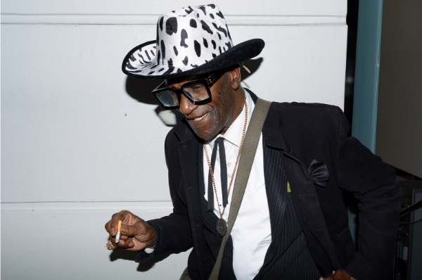 Man in a cowboy hat smiling at the ground with a ciggie in hand